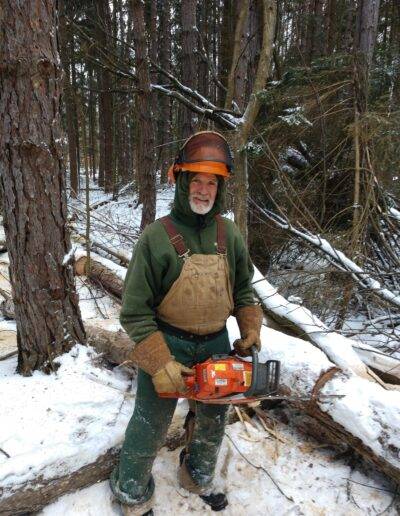 a smiling man stands in snowy woods in wny and holds a chainsaw while trimming trees
